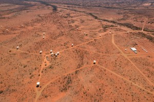 White dish shaped radio telescopes nad a large white building from the air on a red Australian desert backdrop. 