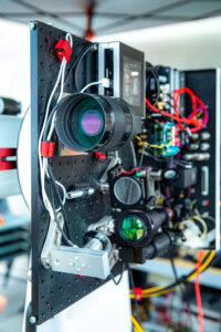 An assembly of lenses, mirrors, cables and lasers are mounted vertically on a panel for testing.